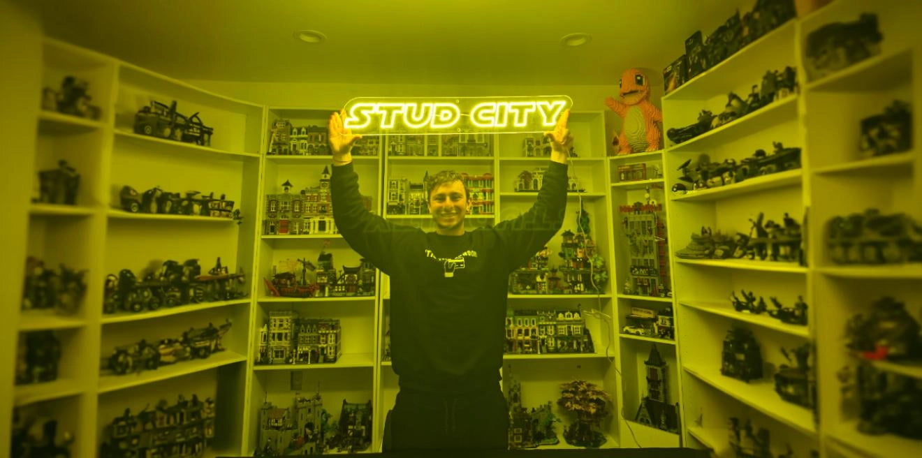 Smiling person standing in a room filled with shelves of LEGO models, holding a glowing neon sign above their head that reads 'STUD CITY'
