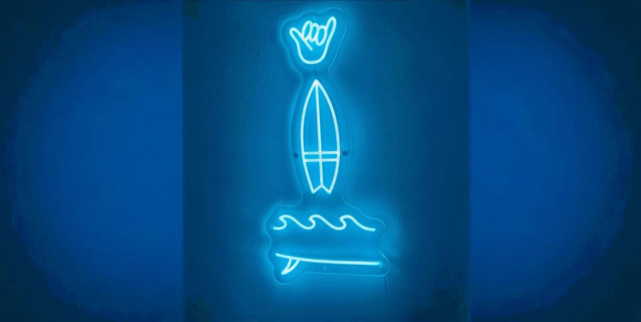 Blue neon sign featuring a shaka hand symbol above a surfboard and ocean wave, glowing against a dark wall.