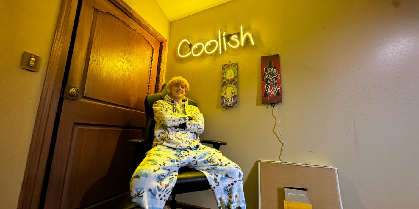 Person in animated pajamas sitting with arms crossed on a chair, posing under a glowing neon sign that reads 'Coolish' next to wall art in a warmly lit room.