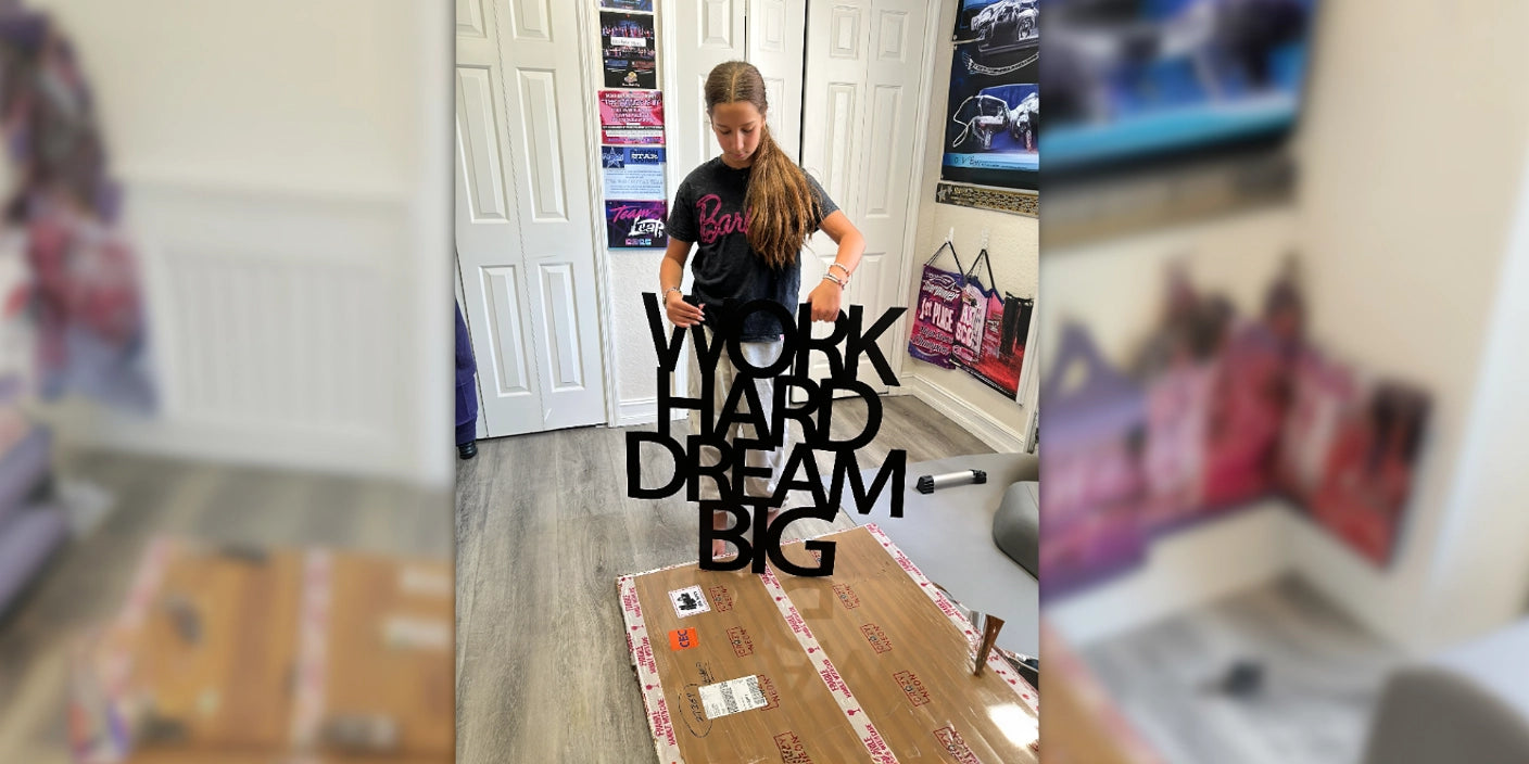 Girl in black Barbie t-shirt holding black metal wall art that says Work Hard Dream Big in trendy home office with posters and packages