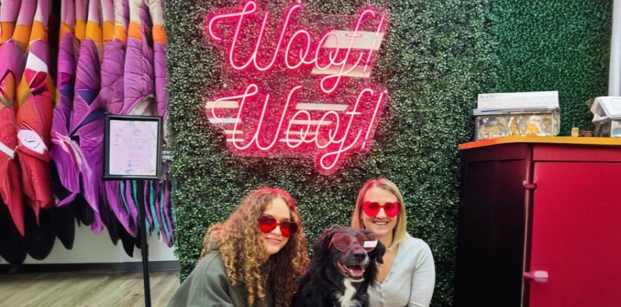 Two women with dog posing in front of pink Woof Woof neon sign at pet boutique with leafy wall
