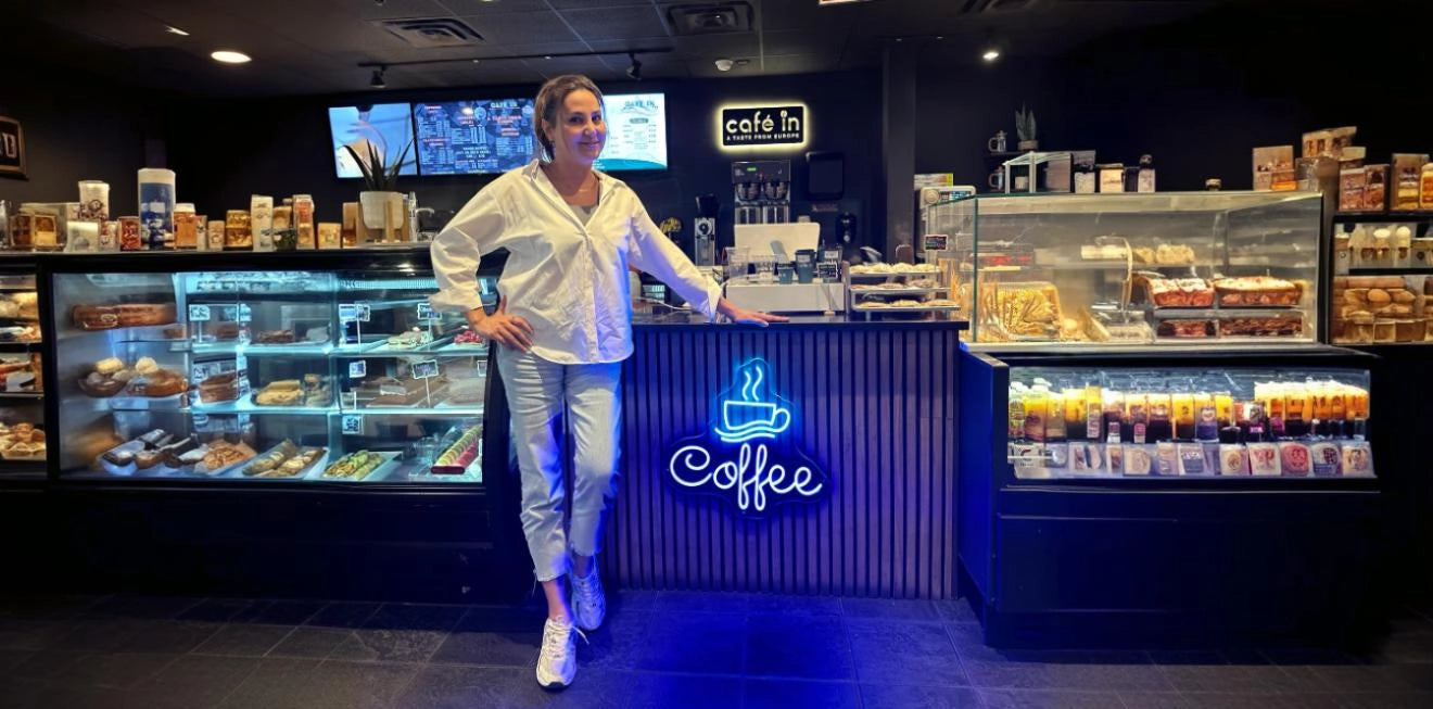 Person standing at the counter of a modern café, with illuminated blue neon coffee cup sign reading 'Coffee', surrounded by pastry and drink display cases in a dark-lit interior.