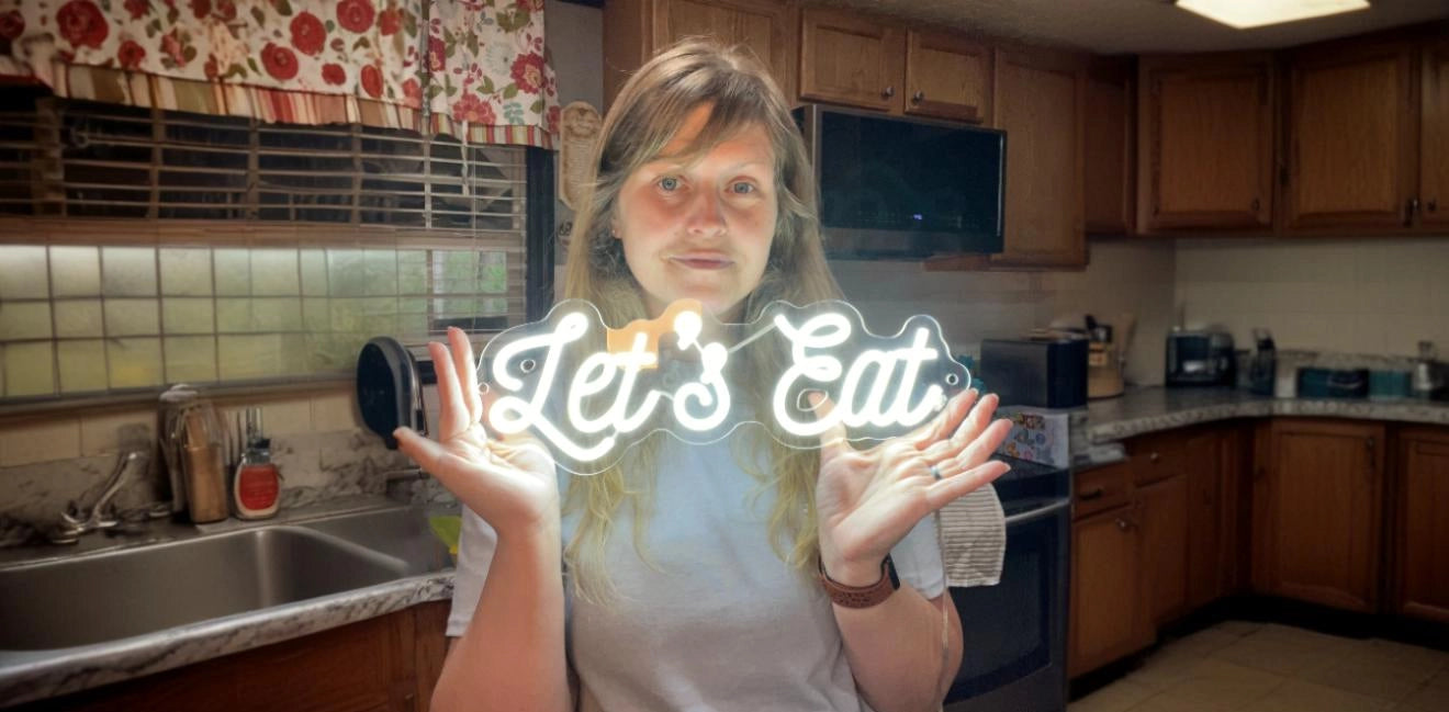 Person standing in a kitchen holding a white neon sign that says 'Let's Eat' in script lettering, with the kitchen background visible