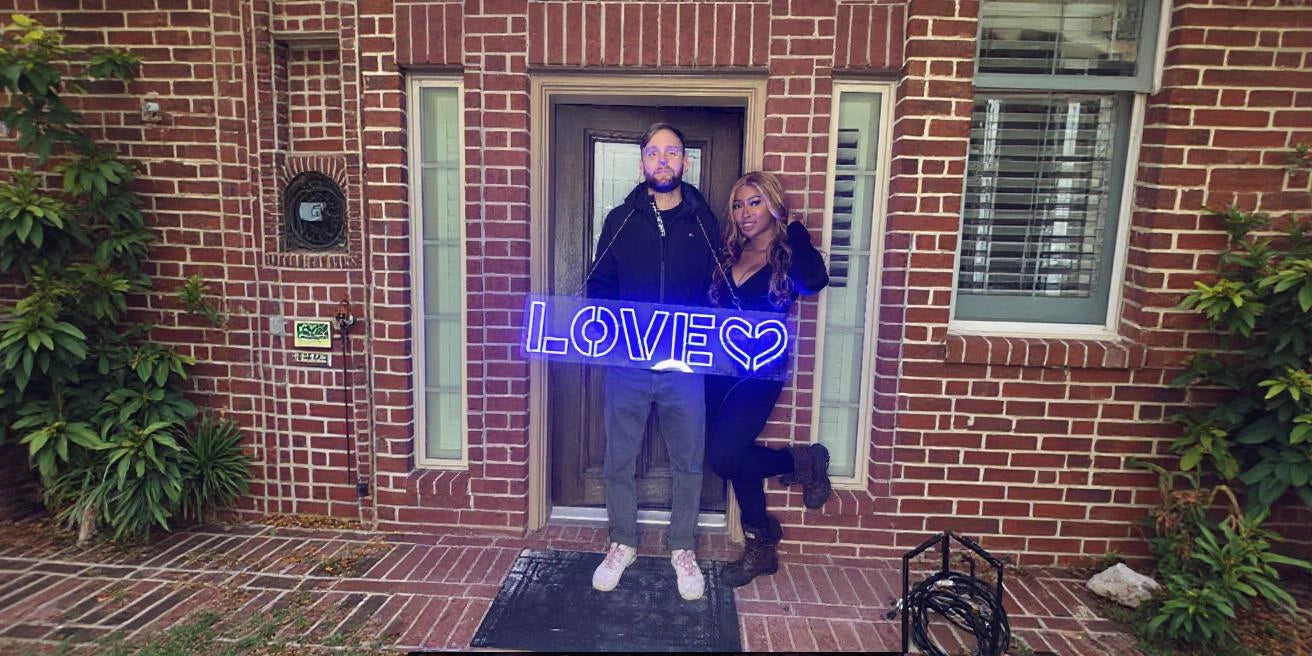 Couple posing in front of brick house entrance holding blue LOVE heart neon sign