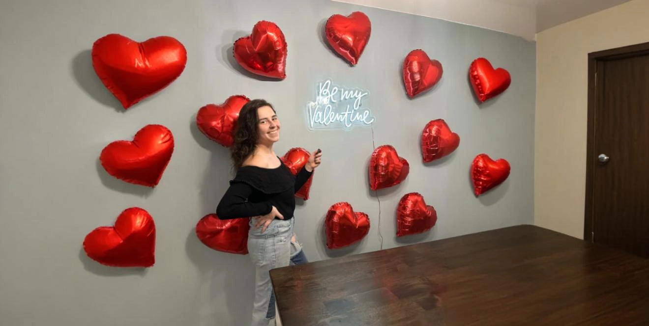 Woman posing with red heart balloons and Be My Valentine white neon sign on gray wall in room