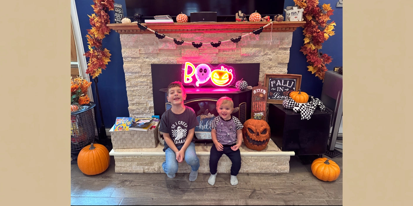 Two children sitting in front of a decorated fireplace with autumn leaves, pumpkins, and a glowing neon sign that reads 'Boo' with Halloween designs