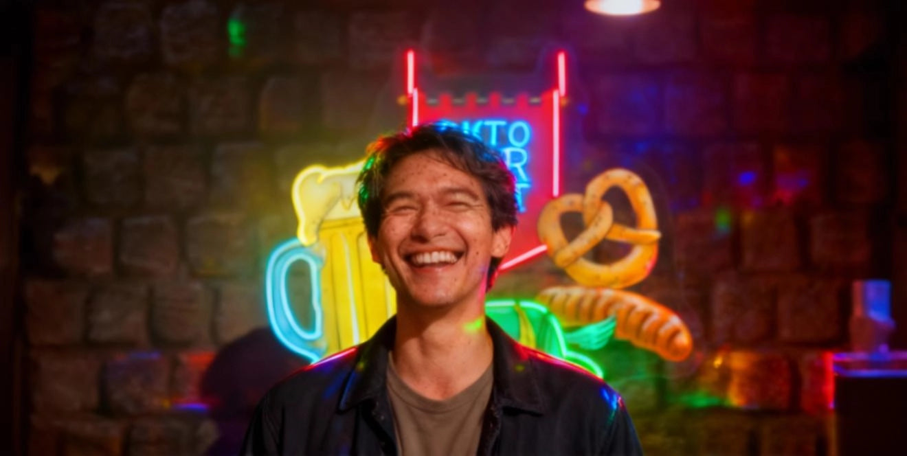 Man standing in front of Oktoberfest-themed neon signs, including a beer mug and pretzel, against a stone wall backdrop