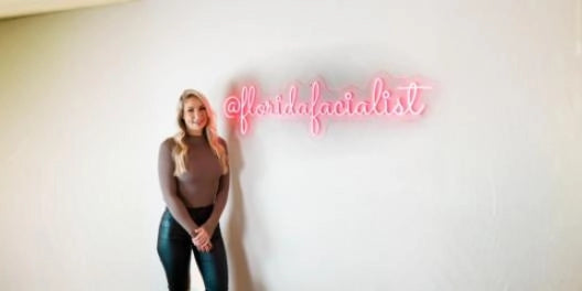 Blonde woman posing beside atfloridafacialist pink neon sign on white wall in minimalist beauty studio