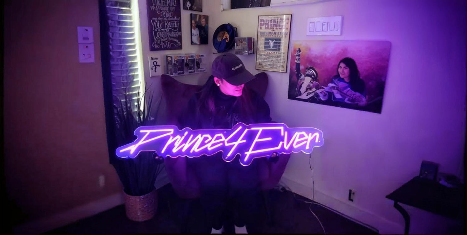 Person sitting in a chair holding a large purple neon sign that reads 'Prince4Ever', surrounded by Prince memorabilia on the walls in a dimly lit room.