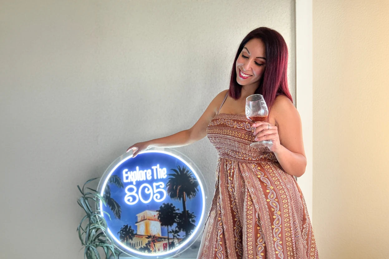 woman in patterned dress holding wine glass standing beside round blue Explore The 805 neon sign in bright coastal lounge with tropical plants and relaxed poolside atmosphere