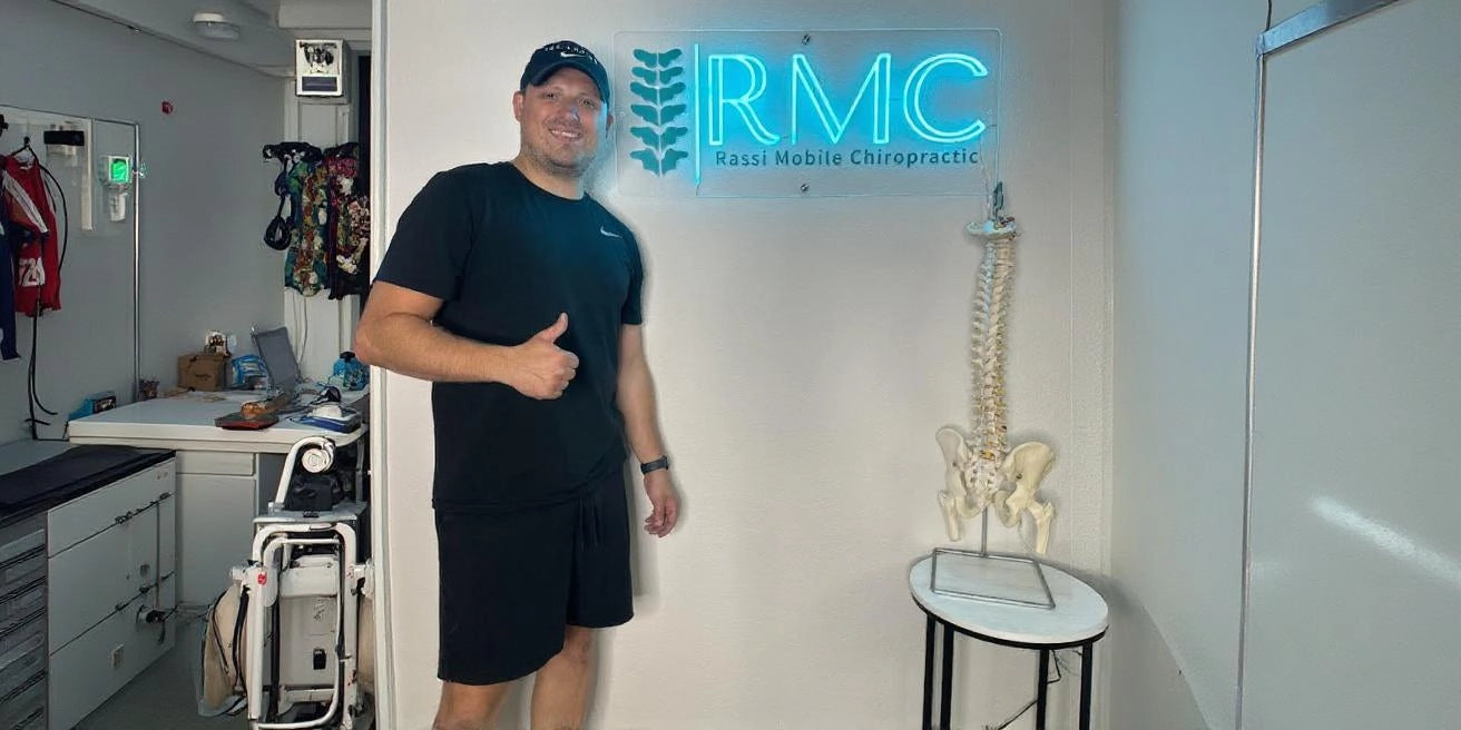 Person in black sportswear giving a thumbs up, standing beside a neon sign that reads 'RMC Rassi Mobile Chiropractic' and a model of a human spine, in a modern clinical setting