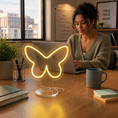 Butterfly Neon Desk Lamp glowing in yellow light on a workspace desk beside a laptop in a modern office.