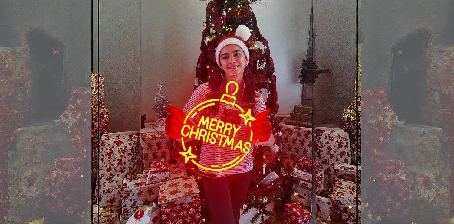 Person in a Santa hat and red striped shirt holding a glowing neon sign that says 'Merry Christmas', standing in front of a large decorated Christmas tree surrounded by wrapped presents