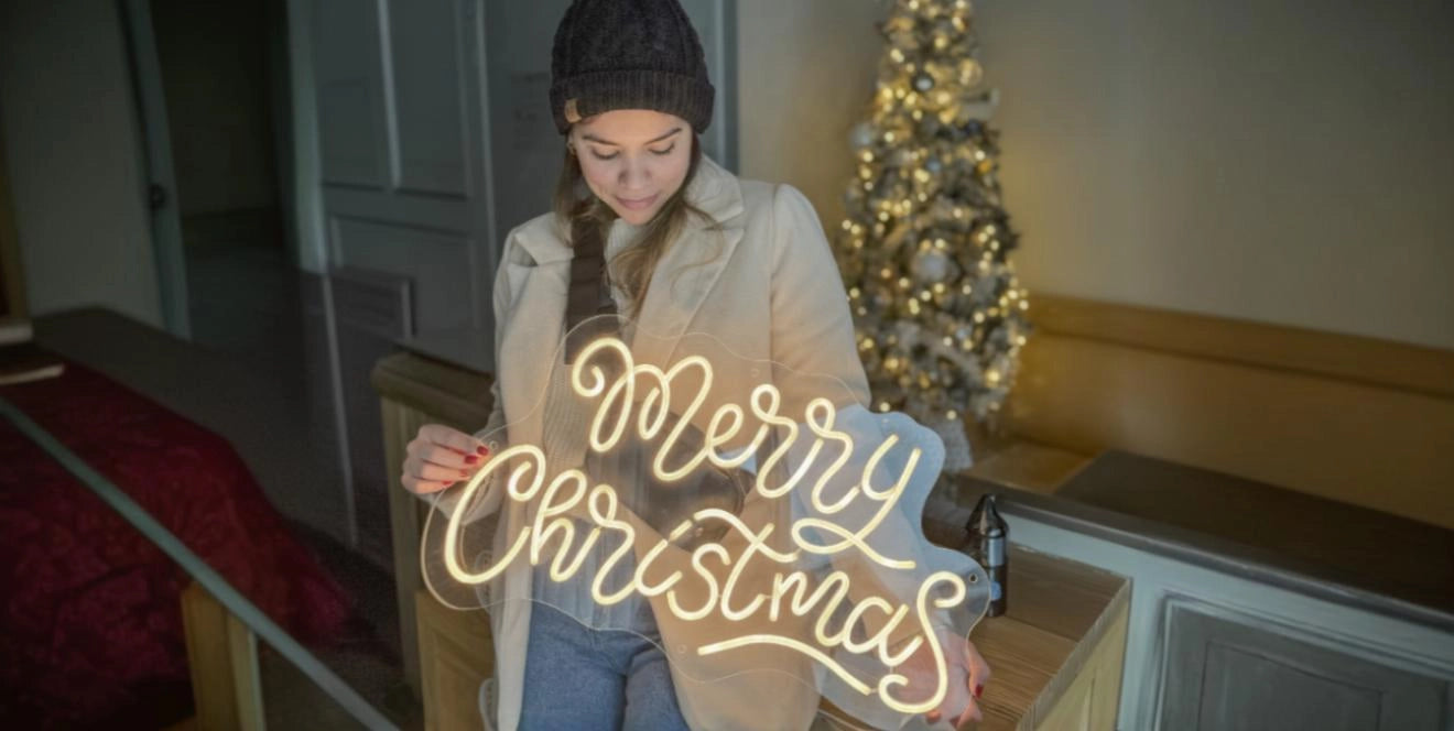 Person in a knit hat and light coat holding a large neon sign that says 'Merry Christmas', standing indoors near a white decorated Christmas tree.