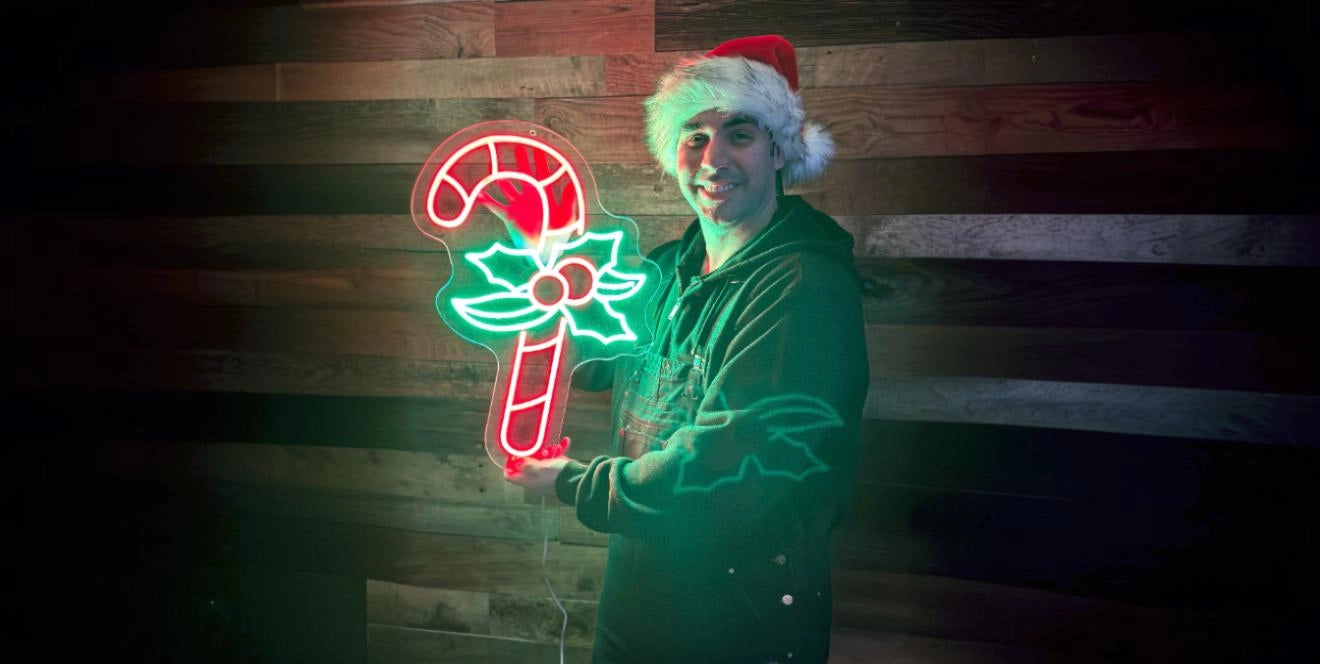 Person wearing a Santa hat and dark hoodie, smiling and holding a colorful neon sign shaped like a candy cane with holly leaves and berries, standing in front of a wooden wall