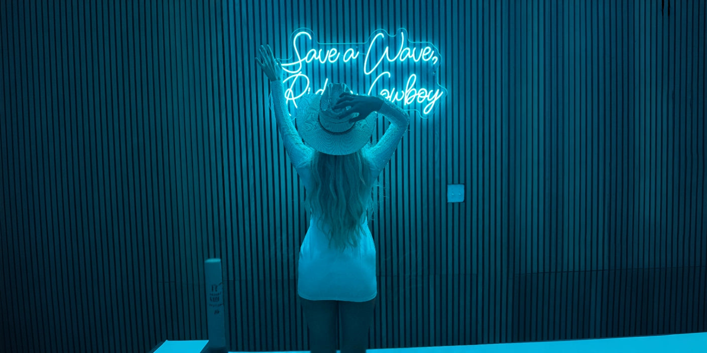 Woman in white dress and cowboy hat standing with arms raised in front of blue Save a Wave Ride a Cowboy neon sign on slatted wall
