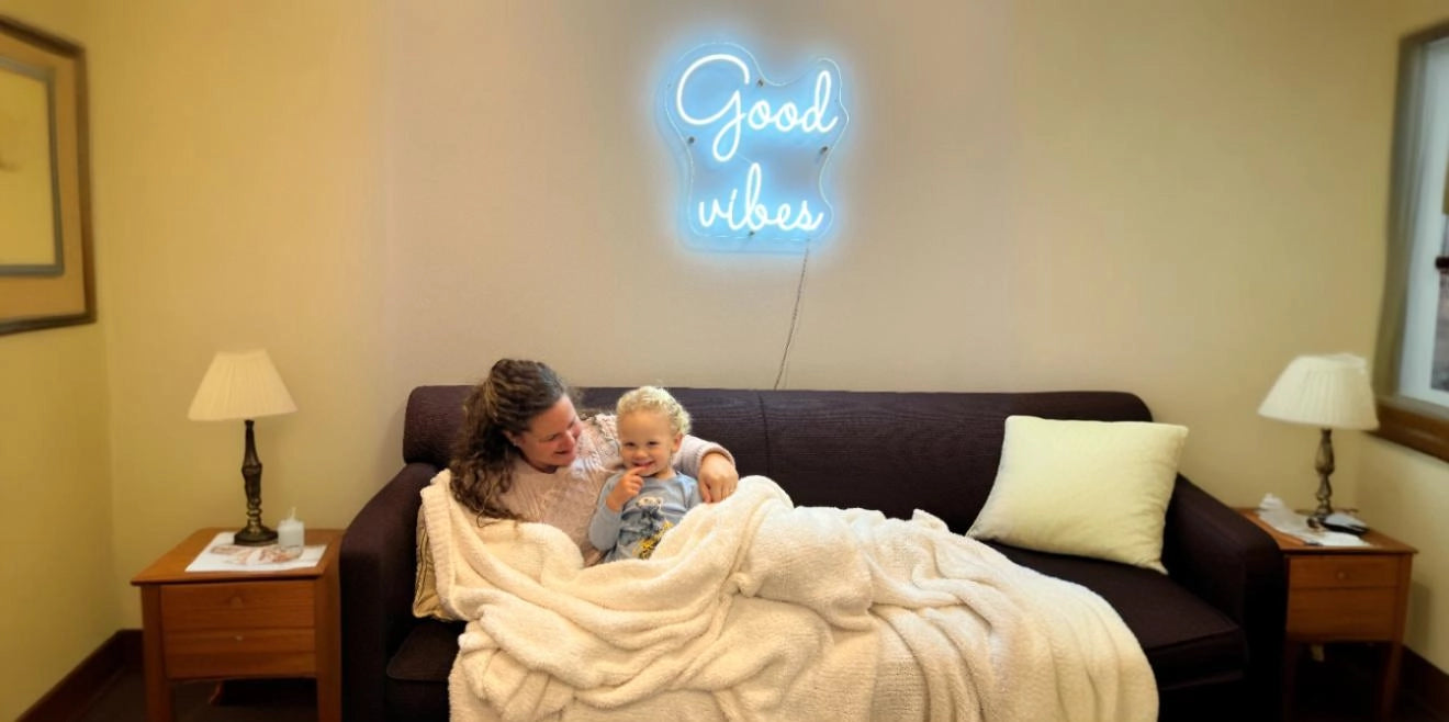 Woman and young child sitting together on a dark couch under a white blanket, smiling beneath a glowing blue neon sign that reads 'Good vibes' in a cozy living room with side tables and lamps