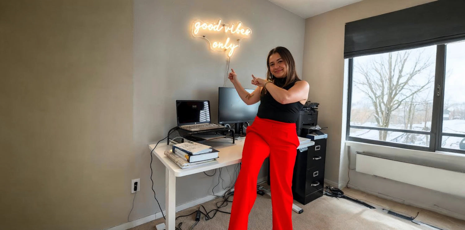 Woman in red pants and black top pointing at a glowing neon sign that reads 'good vibes only', standing beside a computer desk and large window in a modern home office.