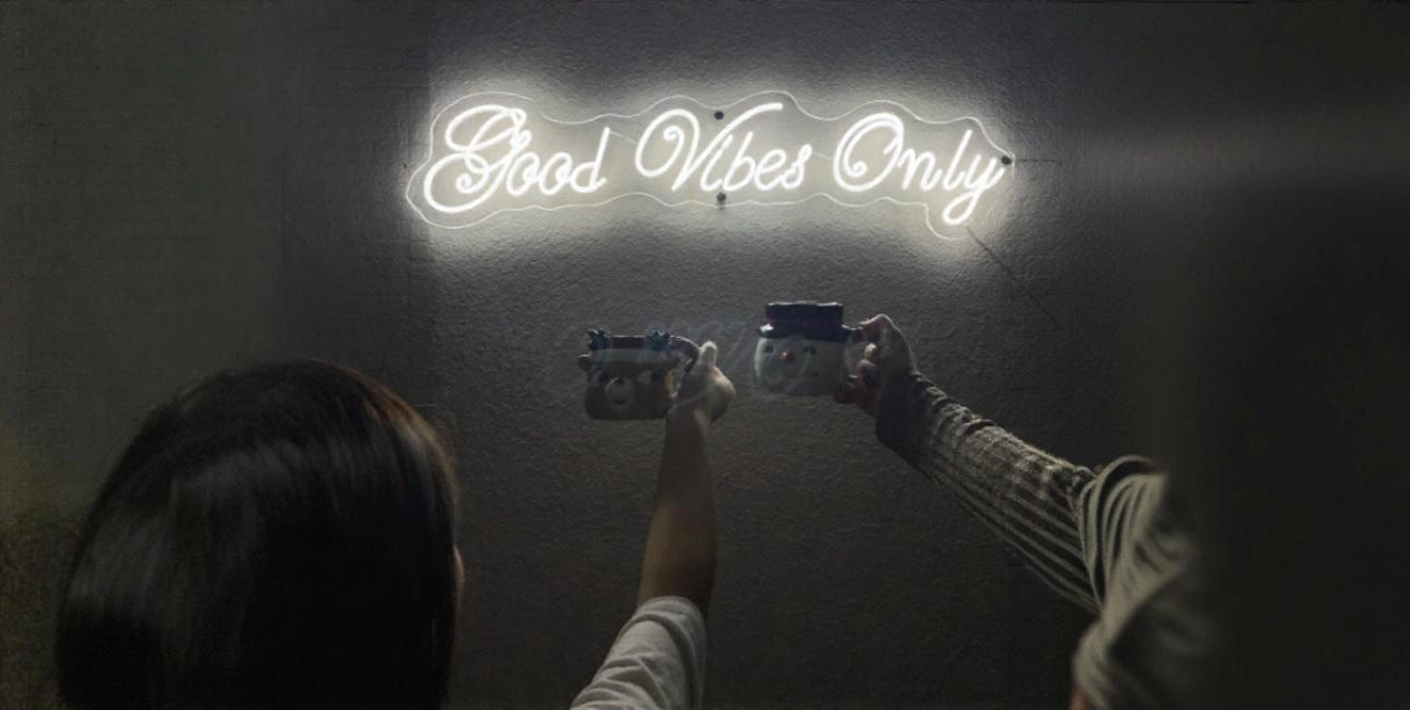 Two people holding up festive mugs and making a toast under a glowing neon sign that reads 'Good Vibes Only' on a dark wall.