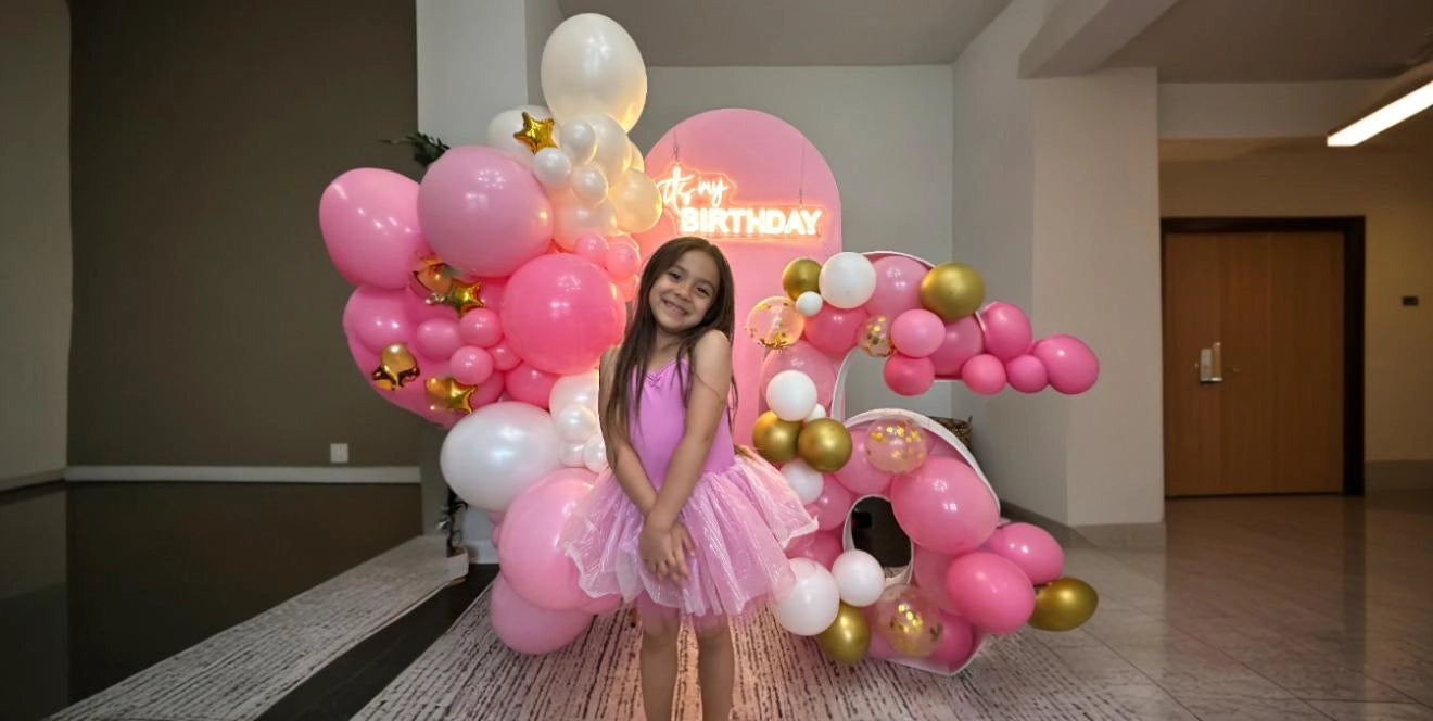 Smiling girl in a pink dress standing in front of a decorative pink, white, and gold balloon arrangement and a glowing neon sign that says 'It's my birthday', in a spacious indoor setting