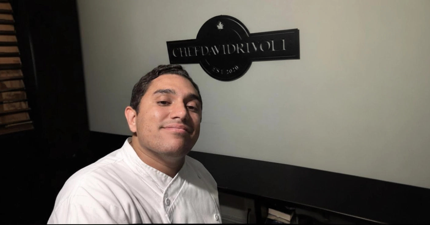 Smiling chef in white coat posing in front of custom black metal monogram Chef David Rivoli Est. 2020 sign on wall in restaurant