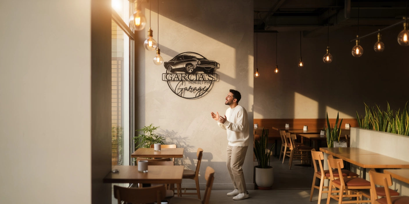 Man in white sweater standing and smiling in sunlit cafe with custom metal monogram Garcias Garage car sign on wall