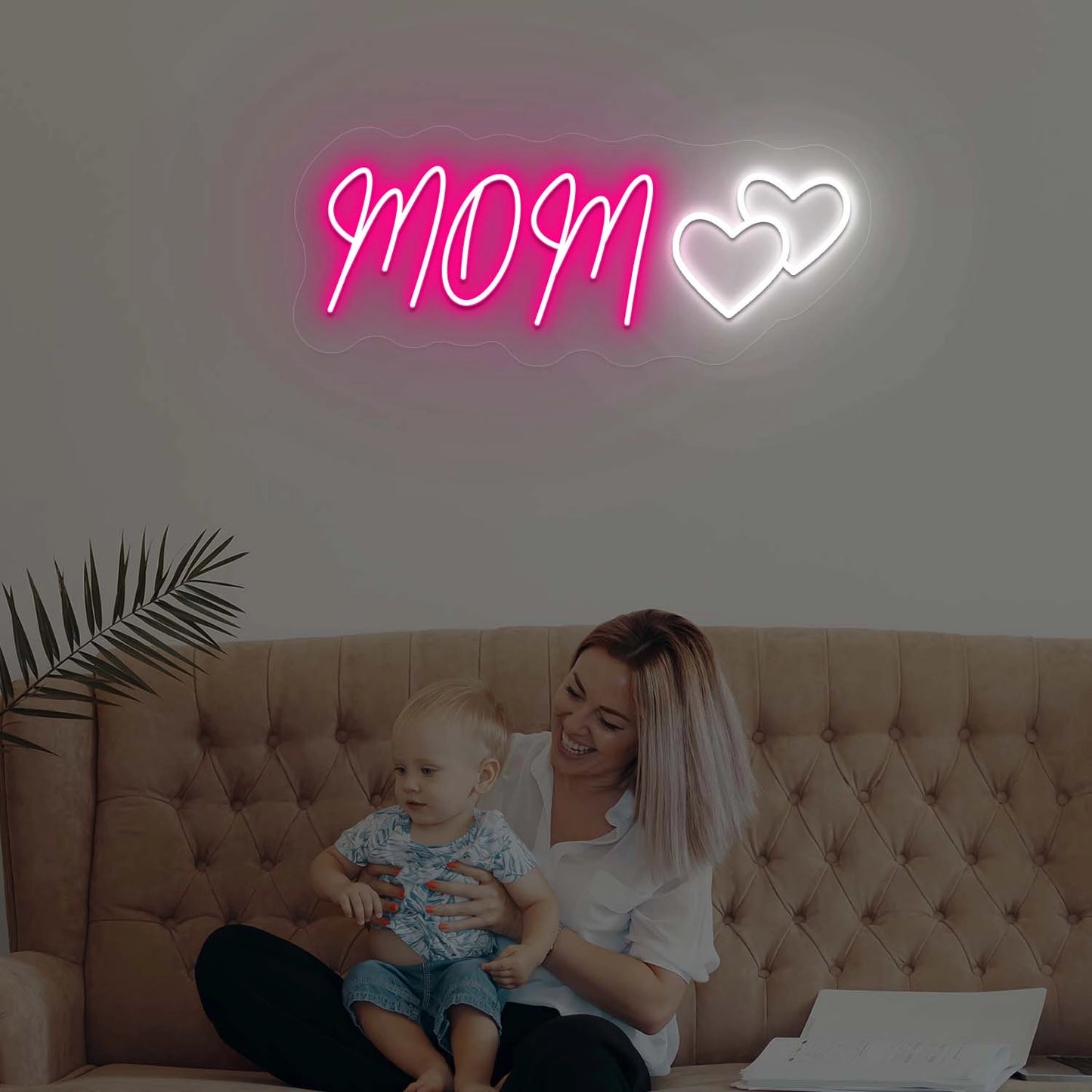 Woman sitting on a couch with a child, illuminated by a neon sign that reads 'MOM' with two hearts.