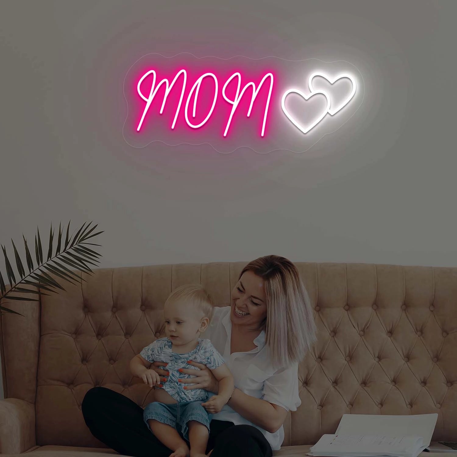 Woman sitting on a couch with a child, illuminated by a neon sign that reads 'MOM' with two hearts.