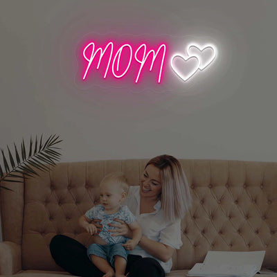 Woman sitting on a couch with a child, illuminated by a neon sign that reads 'MOM' with two hearts.