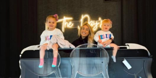 Mom with twins sitting on crib in nursery with The Days Neon Sign glowing in background