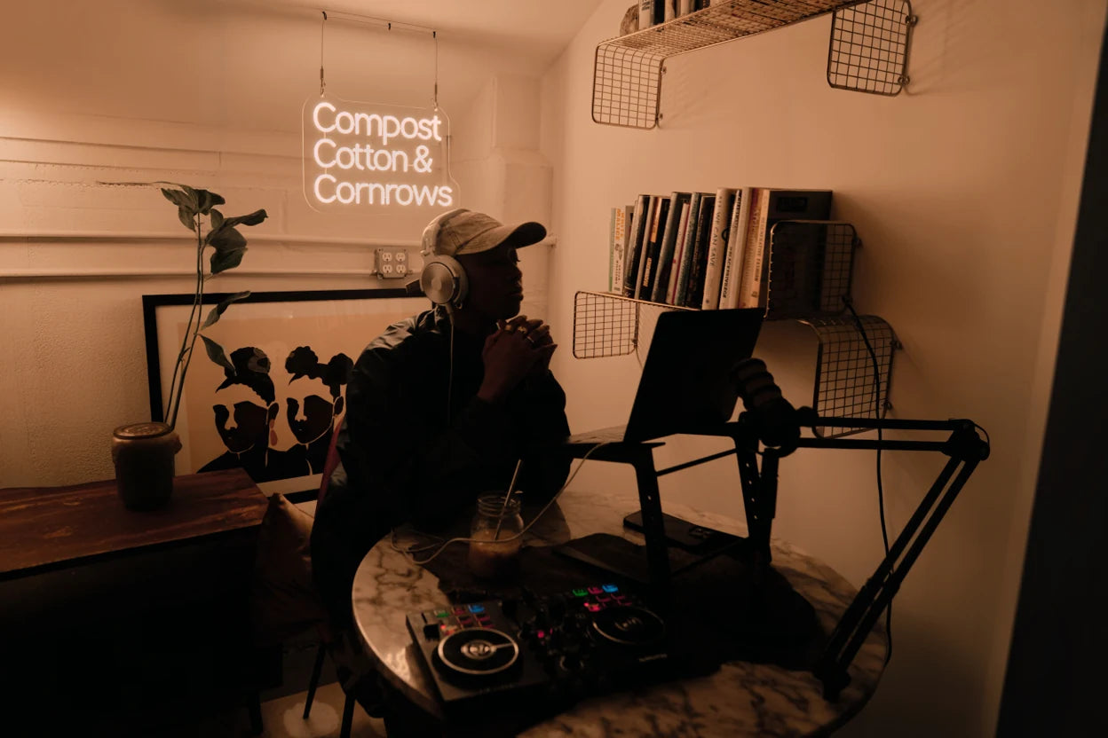 warm lit creative workspace with glowing white custom Compost Cotton and Cornrows neon sign person wearing headphones at desk surrounded by bookshelves laptop DJ controller and framed art