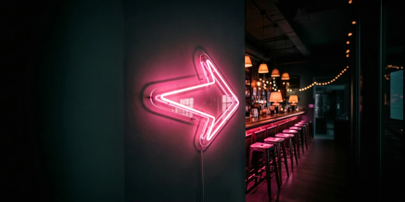 pink right pointing arrow neon sign on dark wall leading toward cozy bar counter with stools hanging lamps and string lights in the background