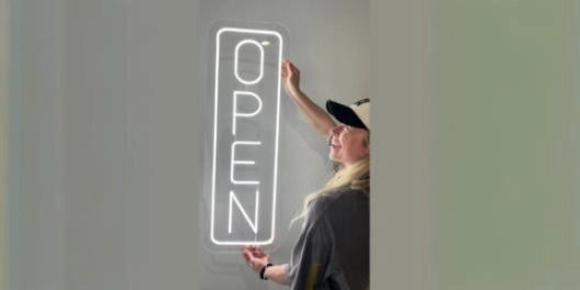 Blonde woman in cap installing vertical white open neon sign on minimalist gray wall
