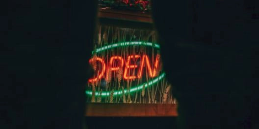 Red and green OPEN neon sign glowing behind window blinds at night, shot through dark curtains