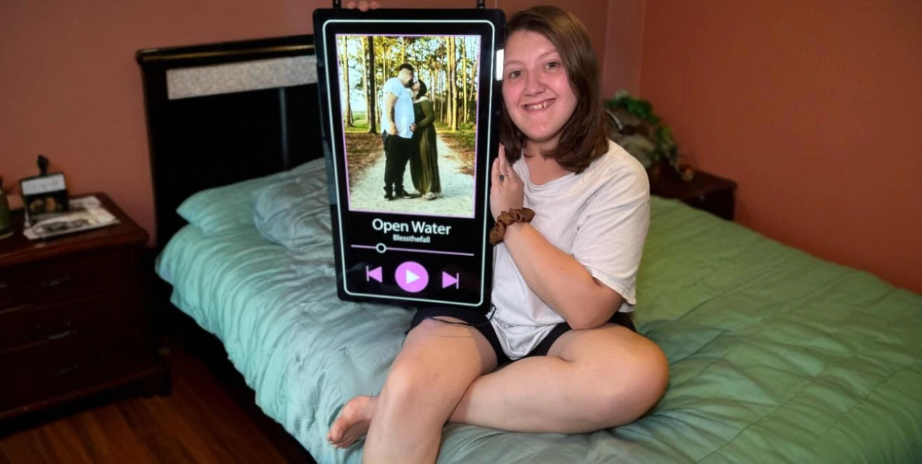Woman in white t-shirt sitting on bed holding large personalized neon artwork designed as music player display with couple photo and Open Water by BlessTheFall song info