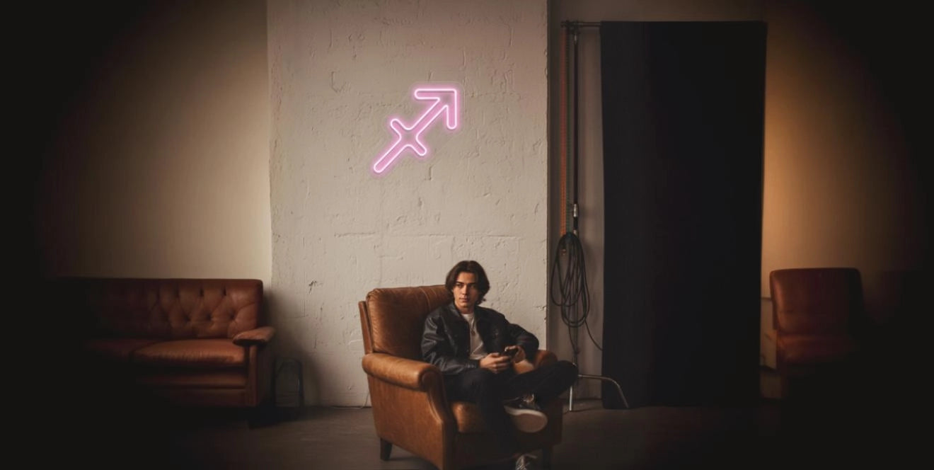 Man in leather jacket sitting on brown armchair under pink Sagittarius zodiac neon sign