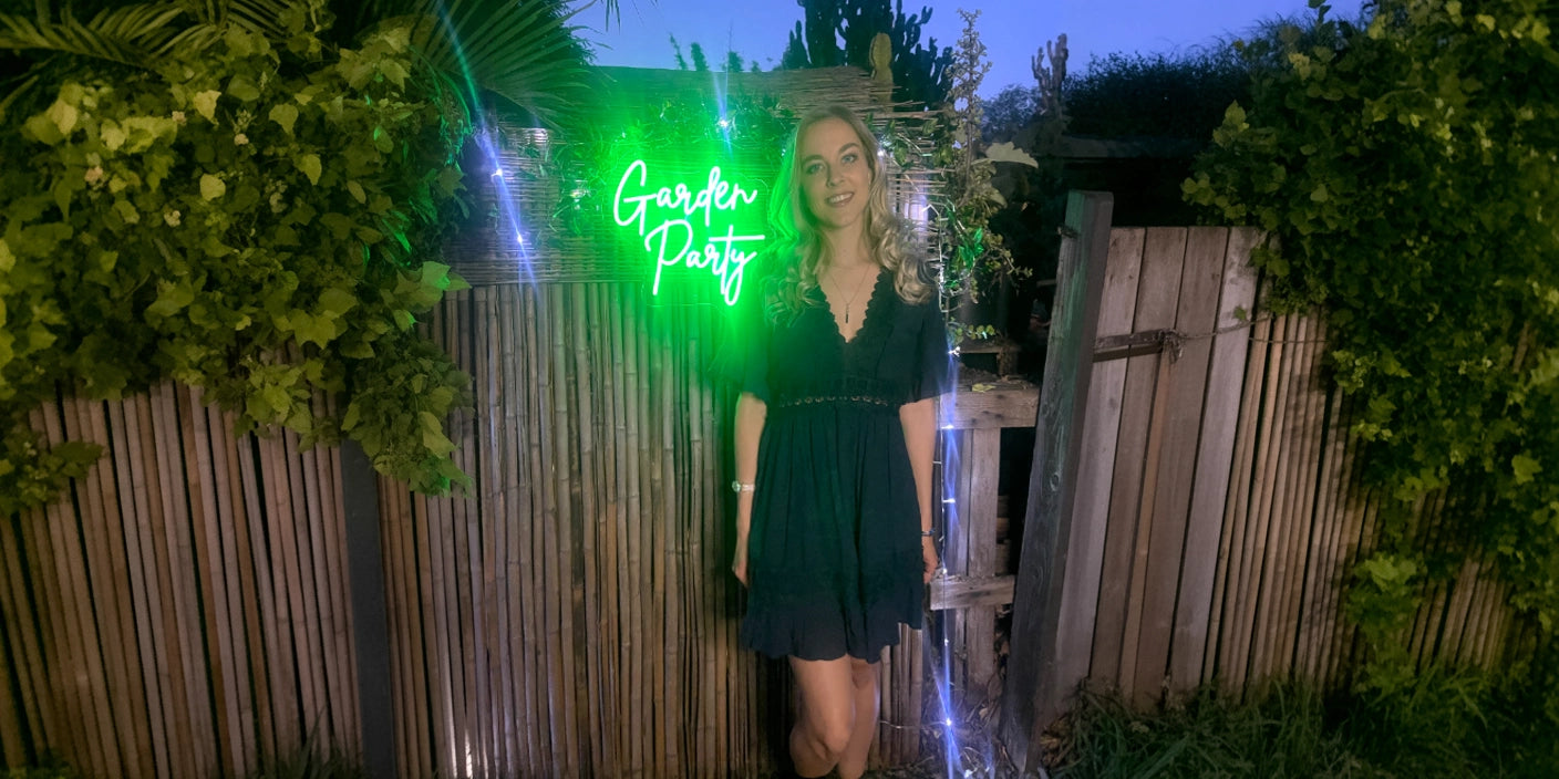 Woman in a dark dress posing beside a wooden garden fence under a glowing green neon sign that reads 'Garden Party' surrounded by lush greenery