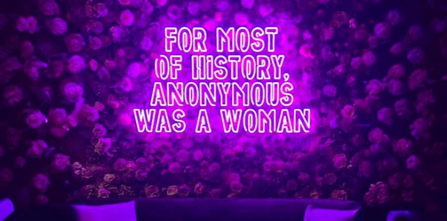 Floral wall with purple For Most of History Anonymous Was a Woman neon quote sign above lounge seating