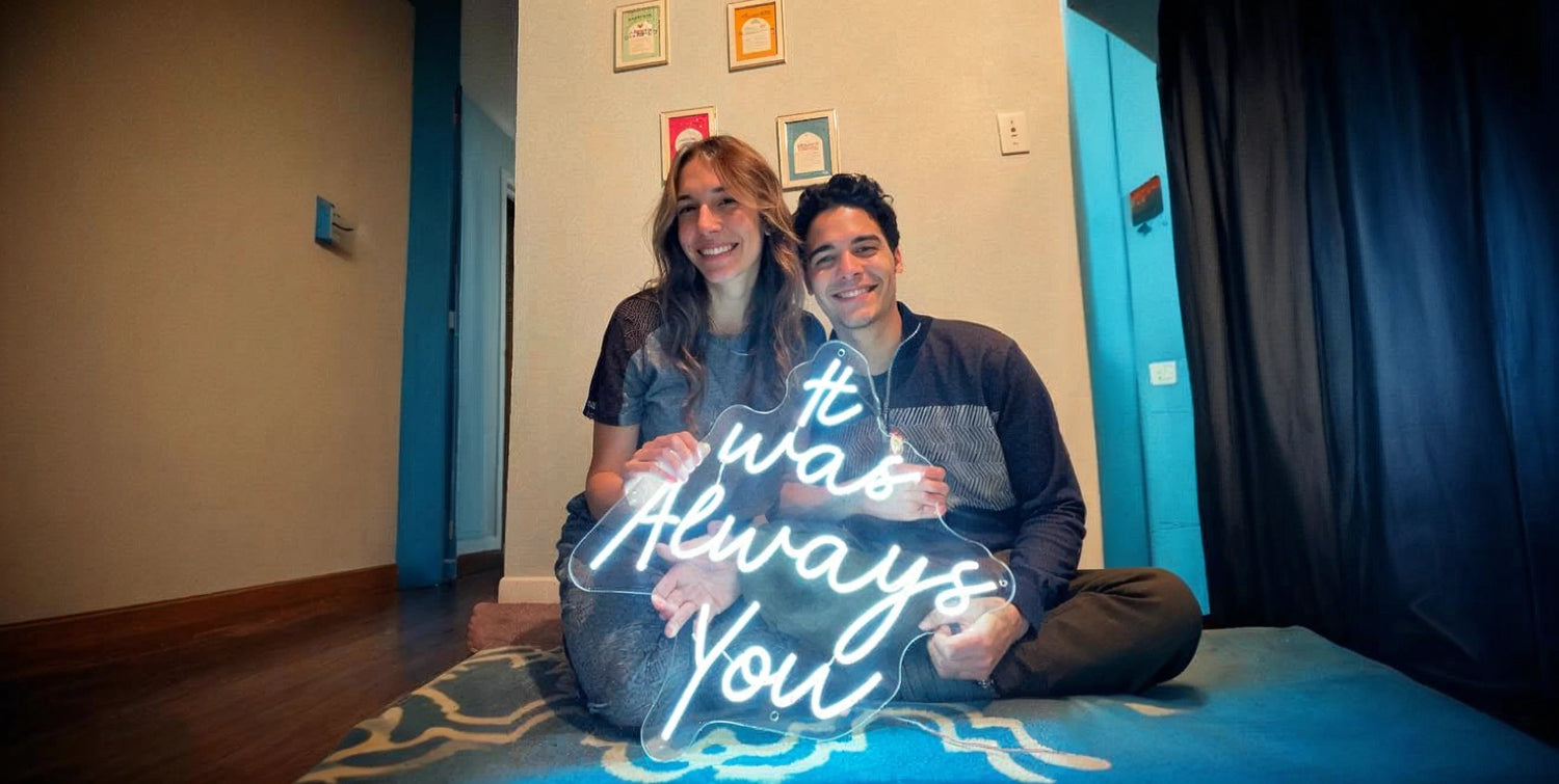 Couple sitting on floor holding It Was Always You white neon sign in cozy room with blue accents and framed artwork